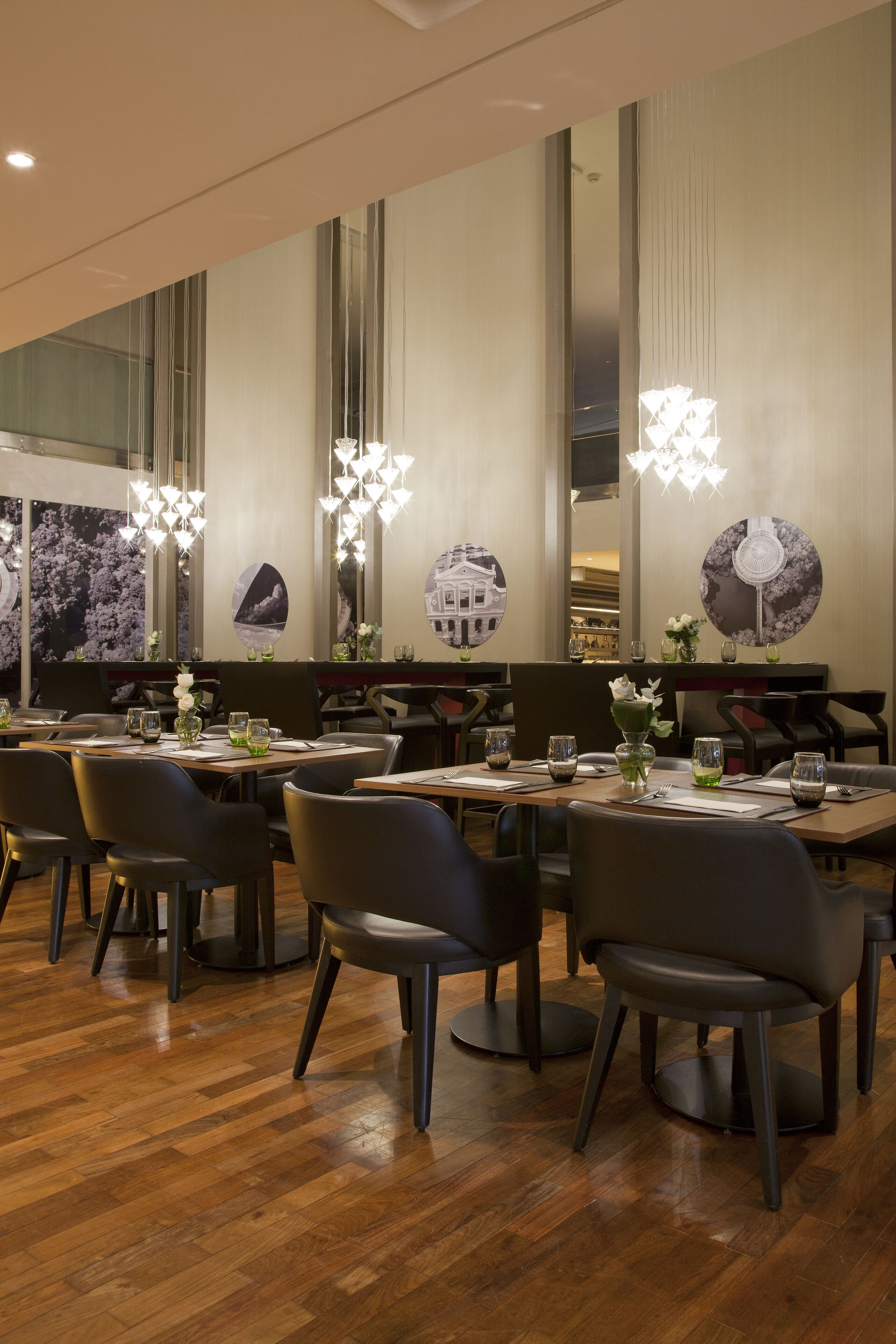 Uma sala de jantar moderna com soalho de madeira, cadeiras pretas e mesas dispostas com copos e centros de mesa florais.