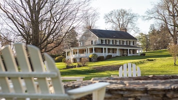 A large house with a porch, surrounded by trees and a grassy hill.