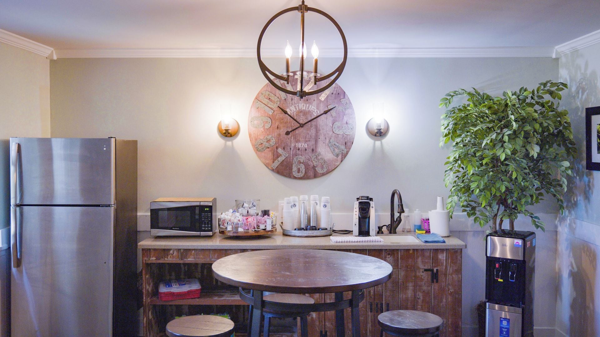 Une cuisine équipée d’un réfrigérateur en acier inoxydable, d’un four à micro-ondes, d’une horloge en bois, d’une table ronde avec des chaises, d’un distributeur d’eau et d’une plante en pot.