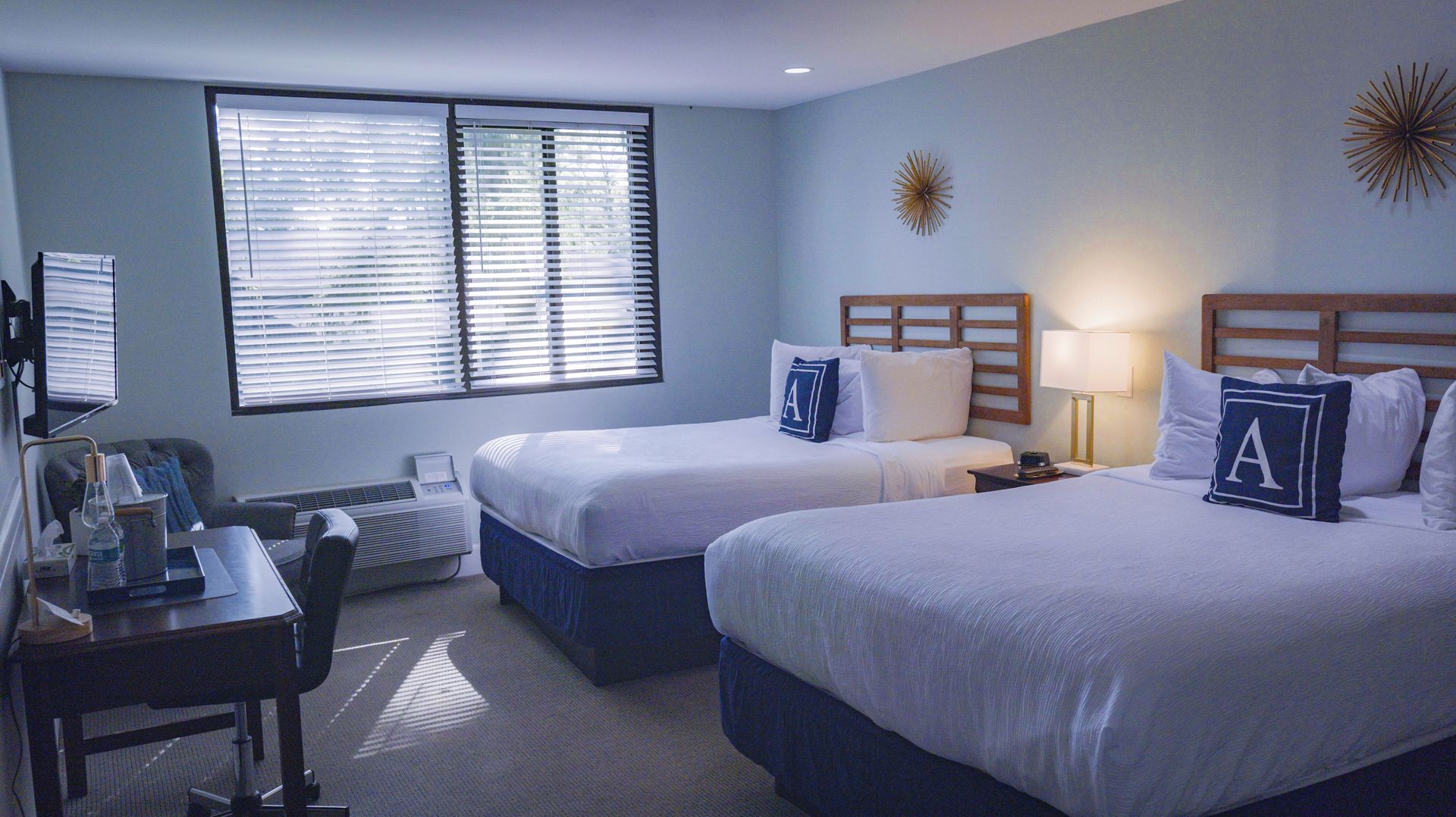 A hotel room with two beds, a desk, and a chair. There is a window with blinds, a wall clock, and a decorative sunburst on the wall.