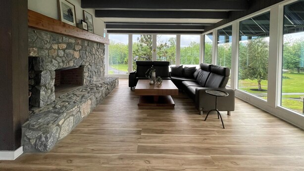 A modern living room with a stone fireplace, a black leather sofa, and large windows offering a view of a green landscape. - Allenberry Resort (Boiling Springs)