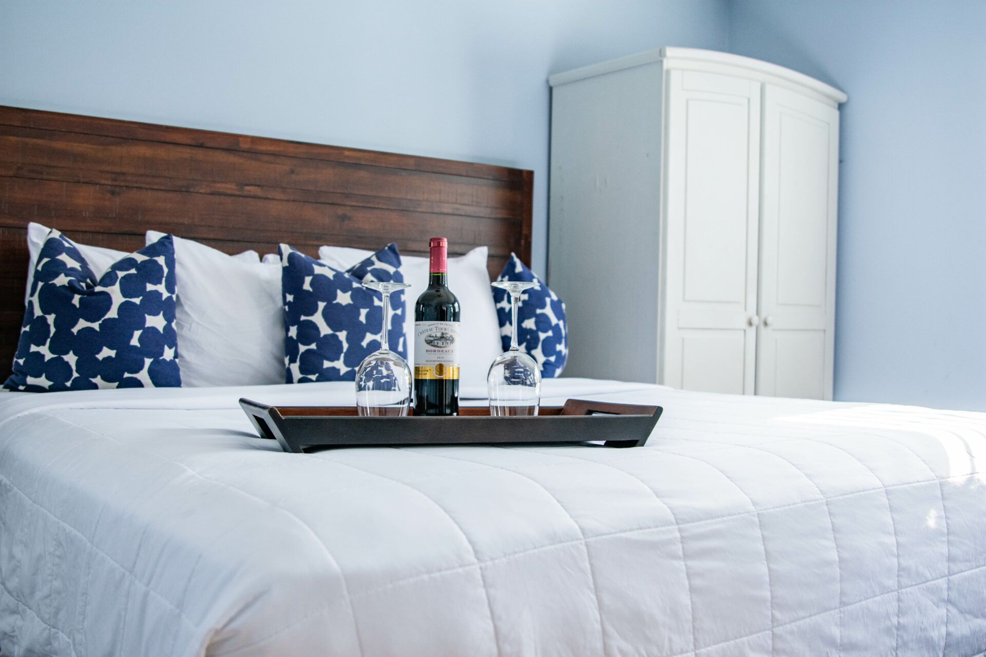 A bed with a tray holding a bottle and two glasses, a wooden headboard, and a white wardrobe.