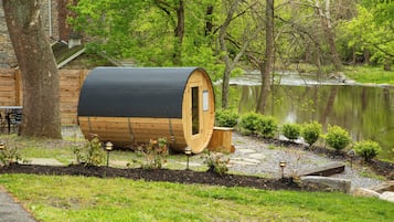 A wooden sauna structure with a door, situated next to a stone building and a body of water, surrounded by greenery.