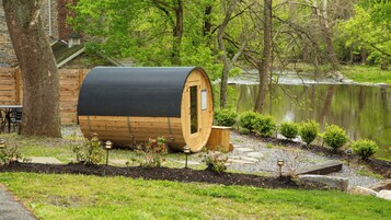 Una estructura de sauna de madera con puerta, ubicada junto a un edificio de piedra y un cuerpo de agua, rodeada de vegetación.