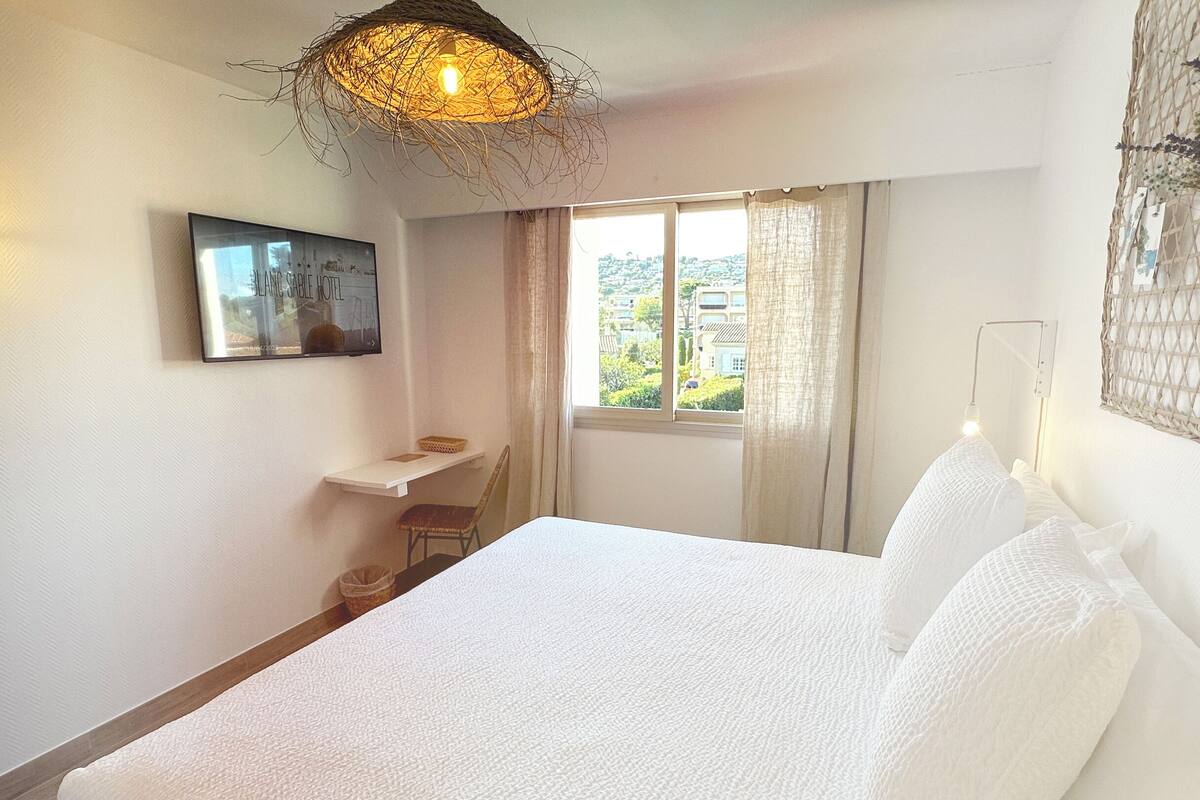 A bedroom with a bed, a chair, a small table, a window with curtains, and a wall-mounted light fixture.