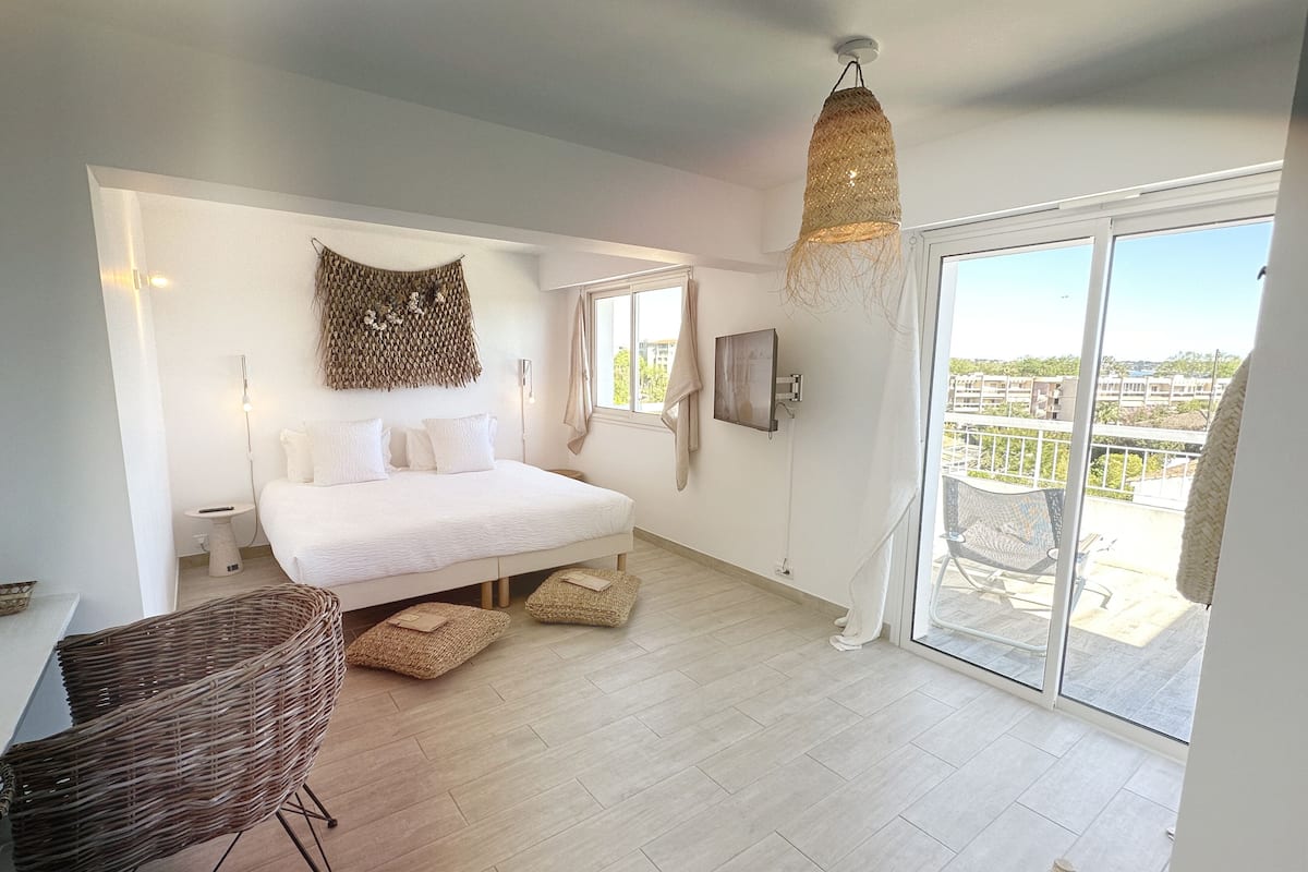 A modern bedroom with a large bed, a wicker chair, a hanging light fixture, and a sliding glass door leading to a balcony.