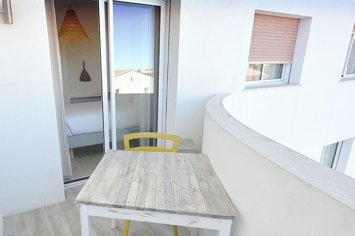 A small balcony with a wooden table and a yellow chair, a sliding glass door leading to a bedroom, and a hanging lamp.