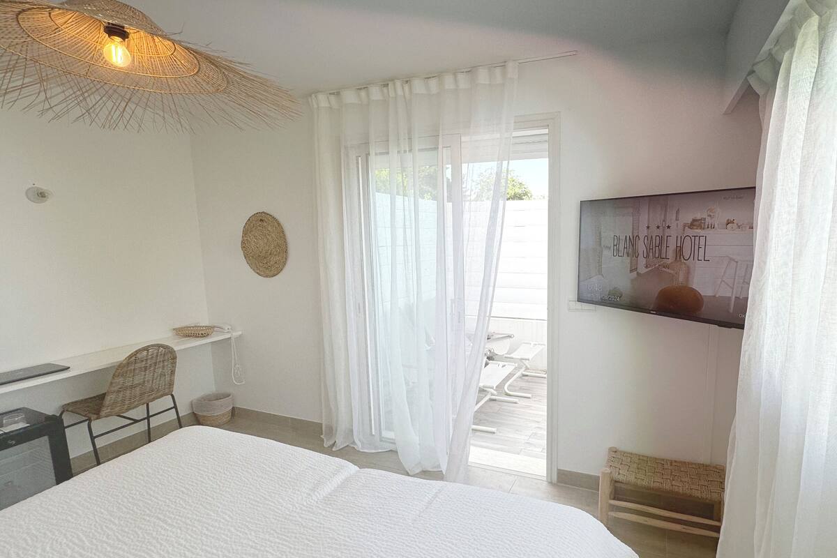 A bedroom with a bed, a desk, a chair, a woven wall hanging, and a view of the outside.