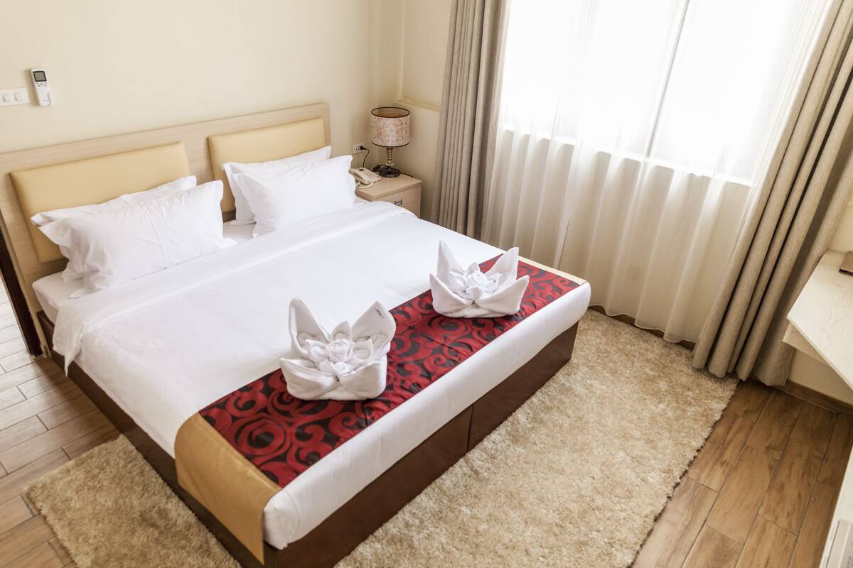 A hotel room with a large bed, a bedside table with a lamp, and a view of the outdoors through the window.