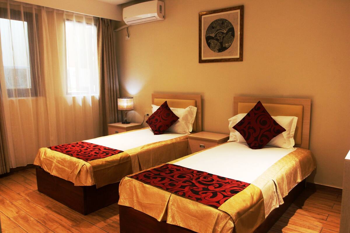 A hotel room with two beds, a nightstand with a lamp, a window with curtains, and an air conditioner on the wall.