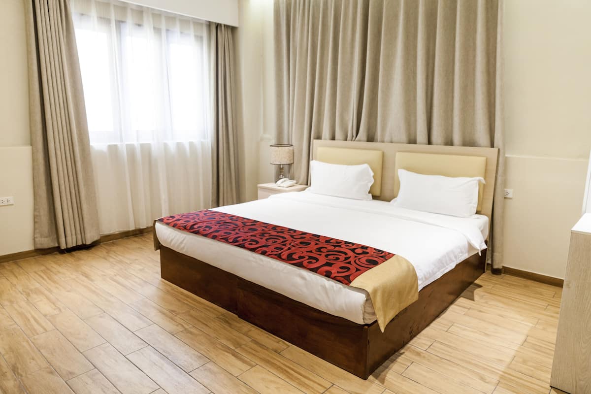 A hotel room with a large bed, wooden flooring, a bedside table with a lamp, and a window with curtains.
