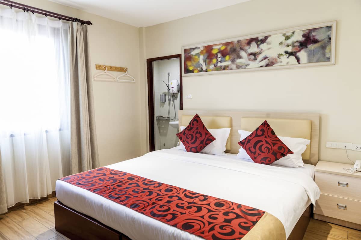 A hotel room with a large bed, a bedside table, a nightstand, a mirror, a shower area, and a painting on the wall.
