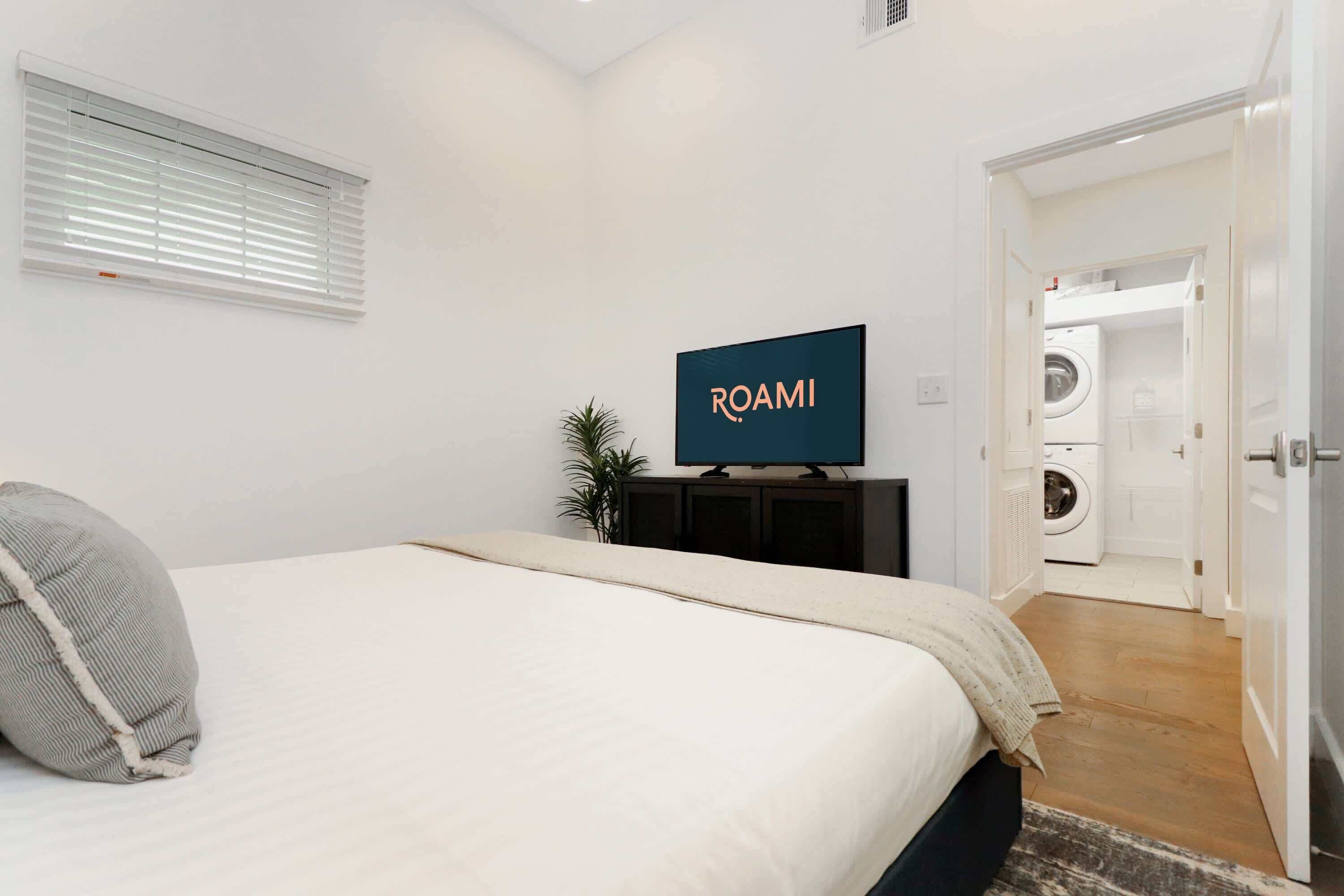 A bedroom with a bed, a television on a stand, a plant, and a laundry room visible through an open door.