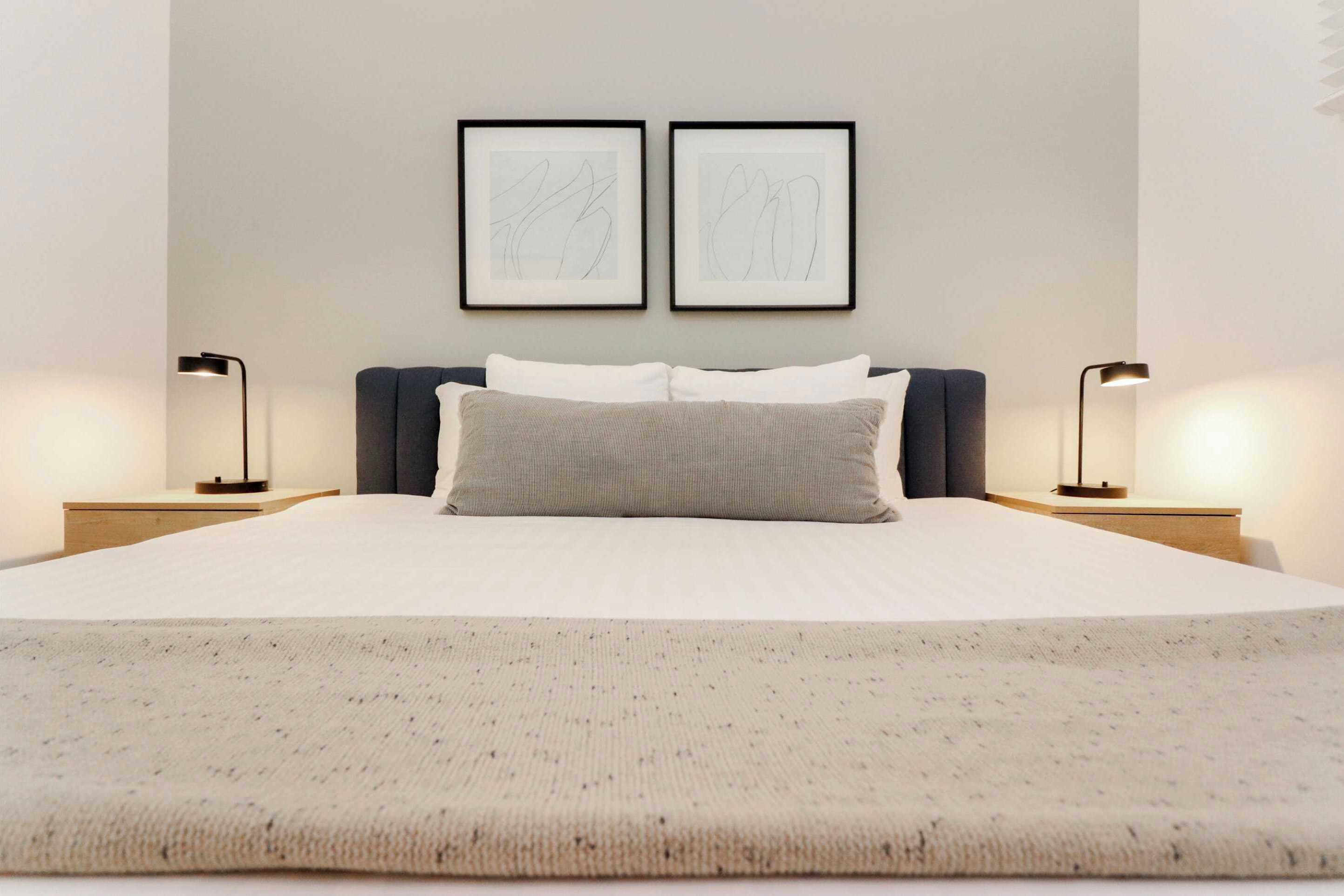 Una cama bien hecha con edredón blanco y dos almohadas, flanqueada por dos mesitas de noche con lámparas, y dos cuadros abstractos enmarcados en la pared de arriba.