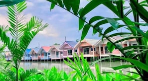 A row of overwater bungalows with a water body in the foreground, surrounded by lush greenery.