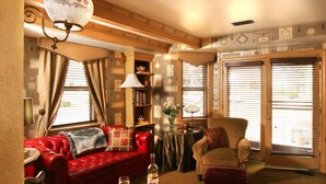 A cozy living room with a red leather sofa, a wooden armchair, and a small table with wine glasses. There is a bookshelf with decorative items and a lamp. - The Queen Victoria B&B (Cape May)