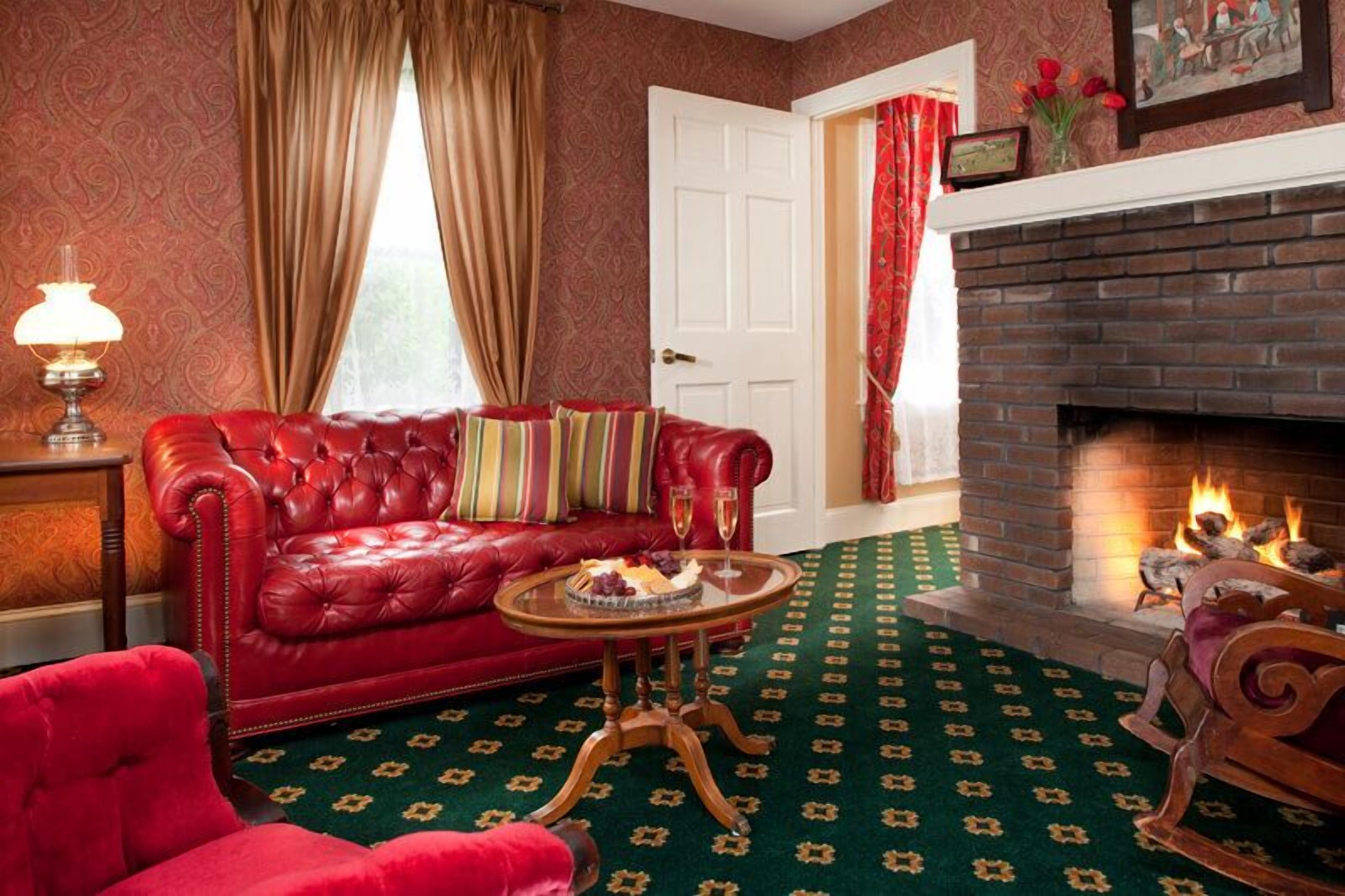 Un canapé en cuir rouge agrémenté de coussins colorés, une petite table ronde avec un panier de fruits et un verre, un tapis à motifs verts et une cheminée en briques où le feu crépite.