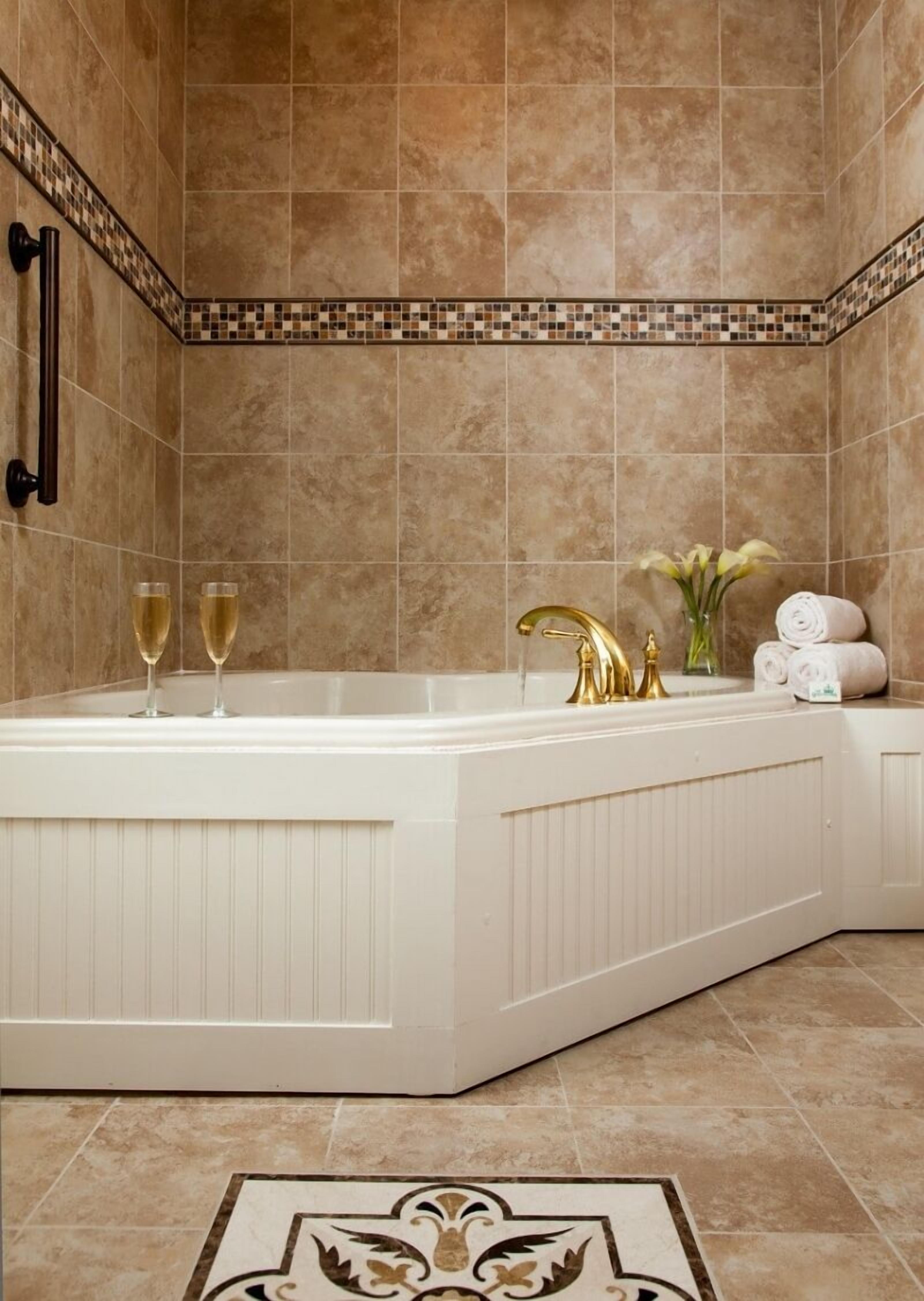 A bathroom with a large bathtub, two wine glasses, a gold faucet, and a tiled backsplash.