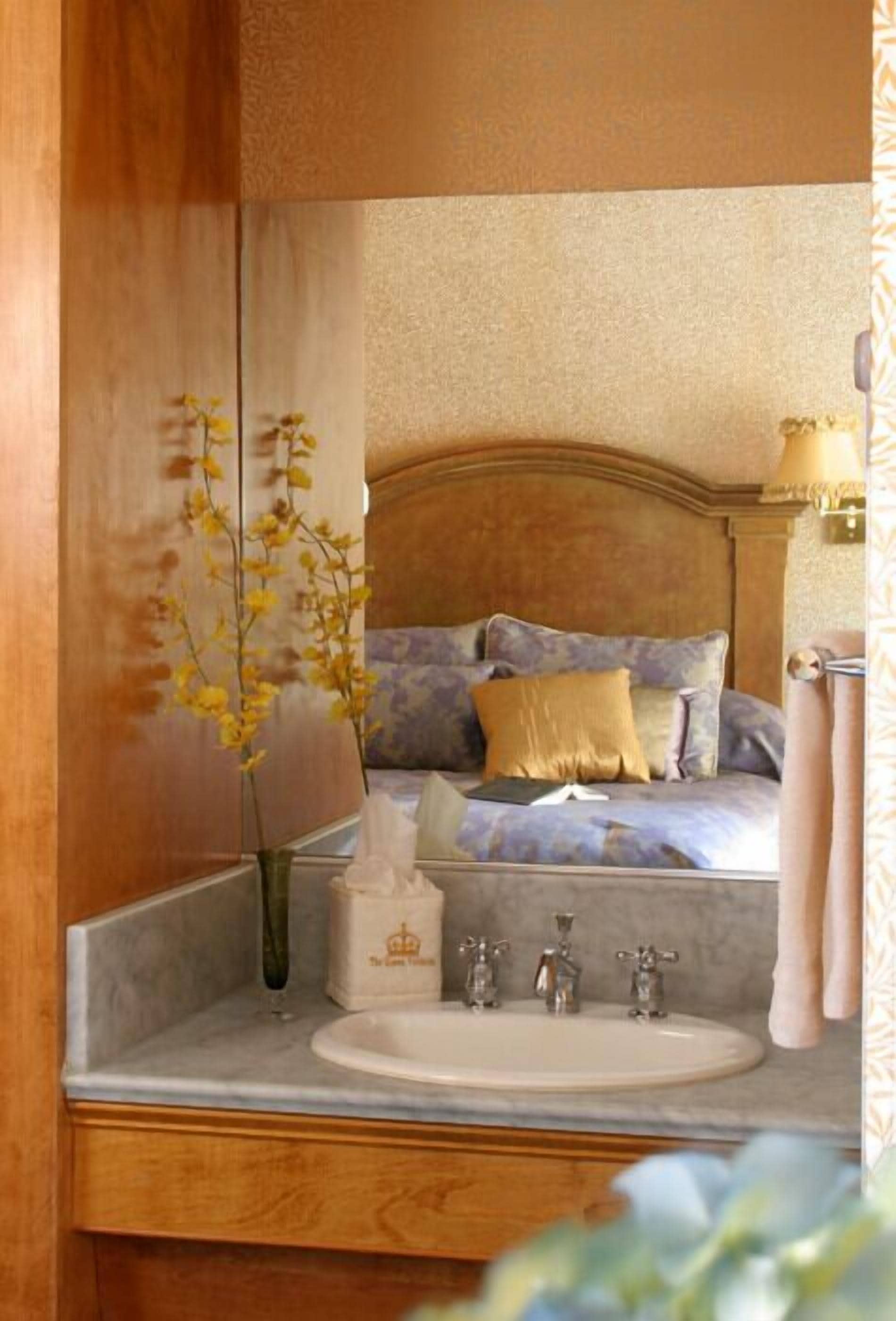 Een slaapkamer met een bed, een nachtkastje met een bloevas, een spiegel en een badkamer met een wastafel en handdoekhouder.