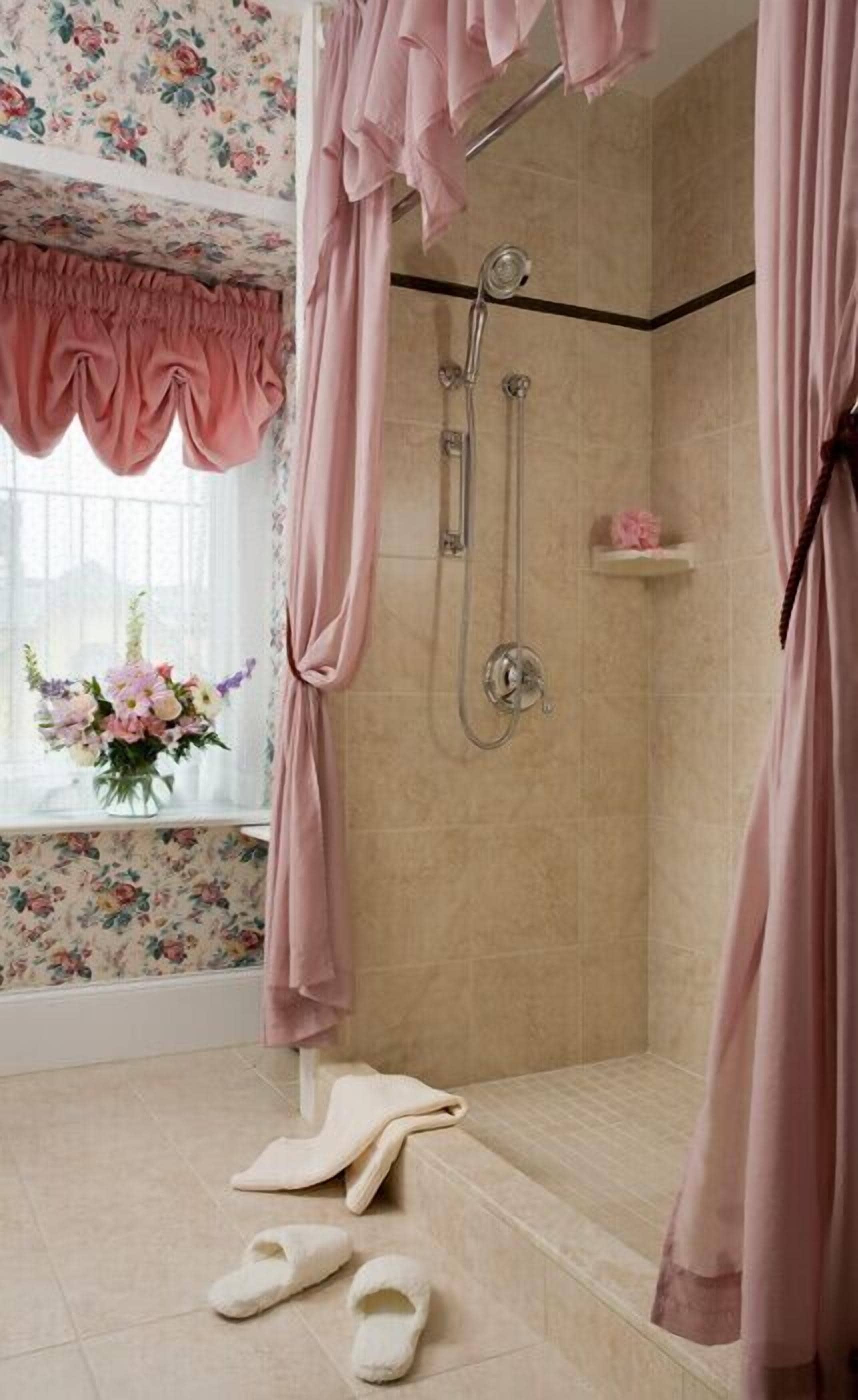 Un espace douche doté d’une porte en verre, une fenêtre ornée de rideaux à motifs floraux et un vase de fleurs posé sur le rebord de la fenêtre.