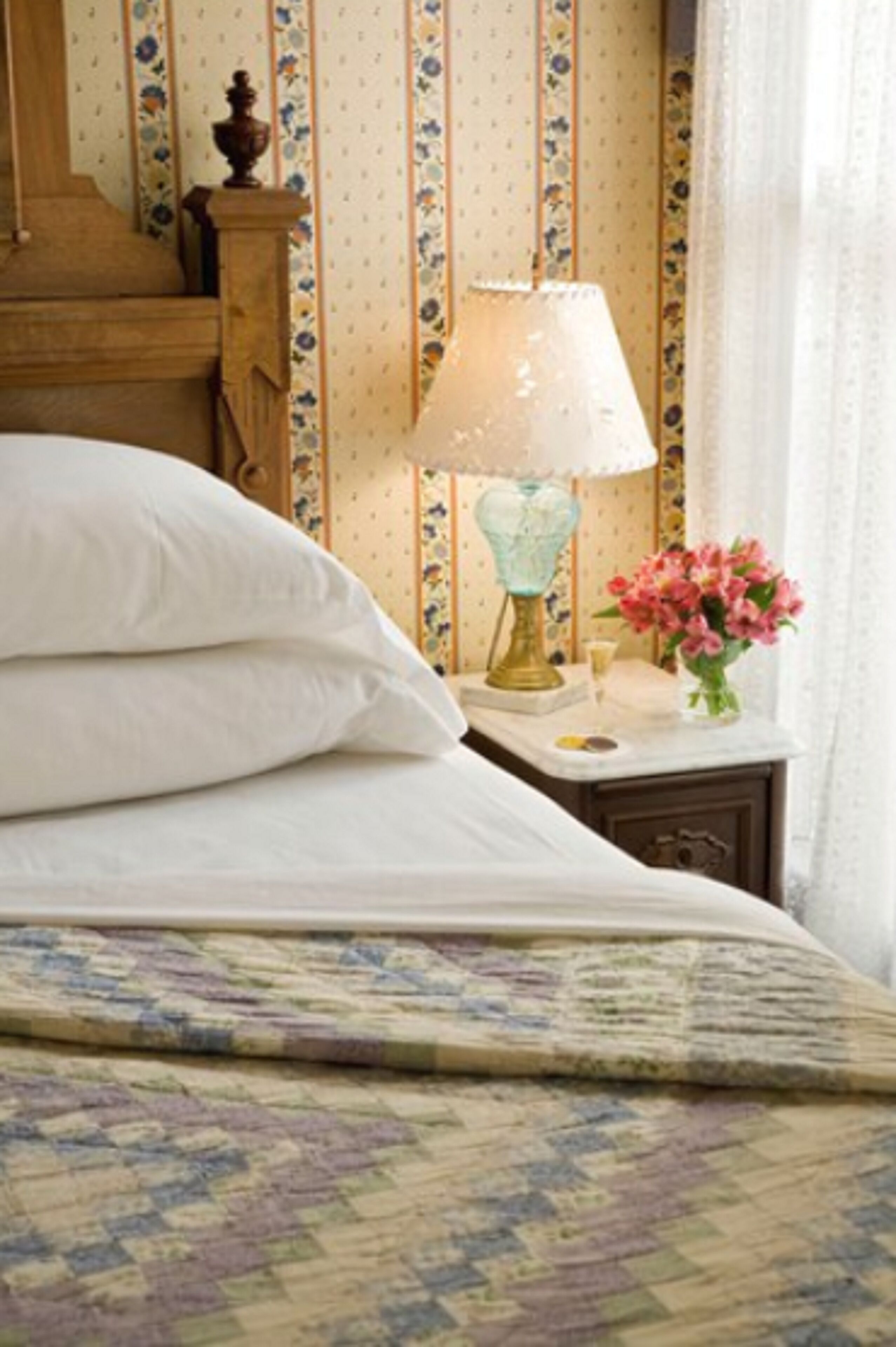 A neatly made bed with white linens, a patterned bedspread, a bedside table with a lamp, and a vase of flowers.