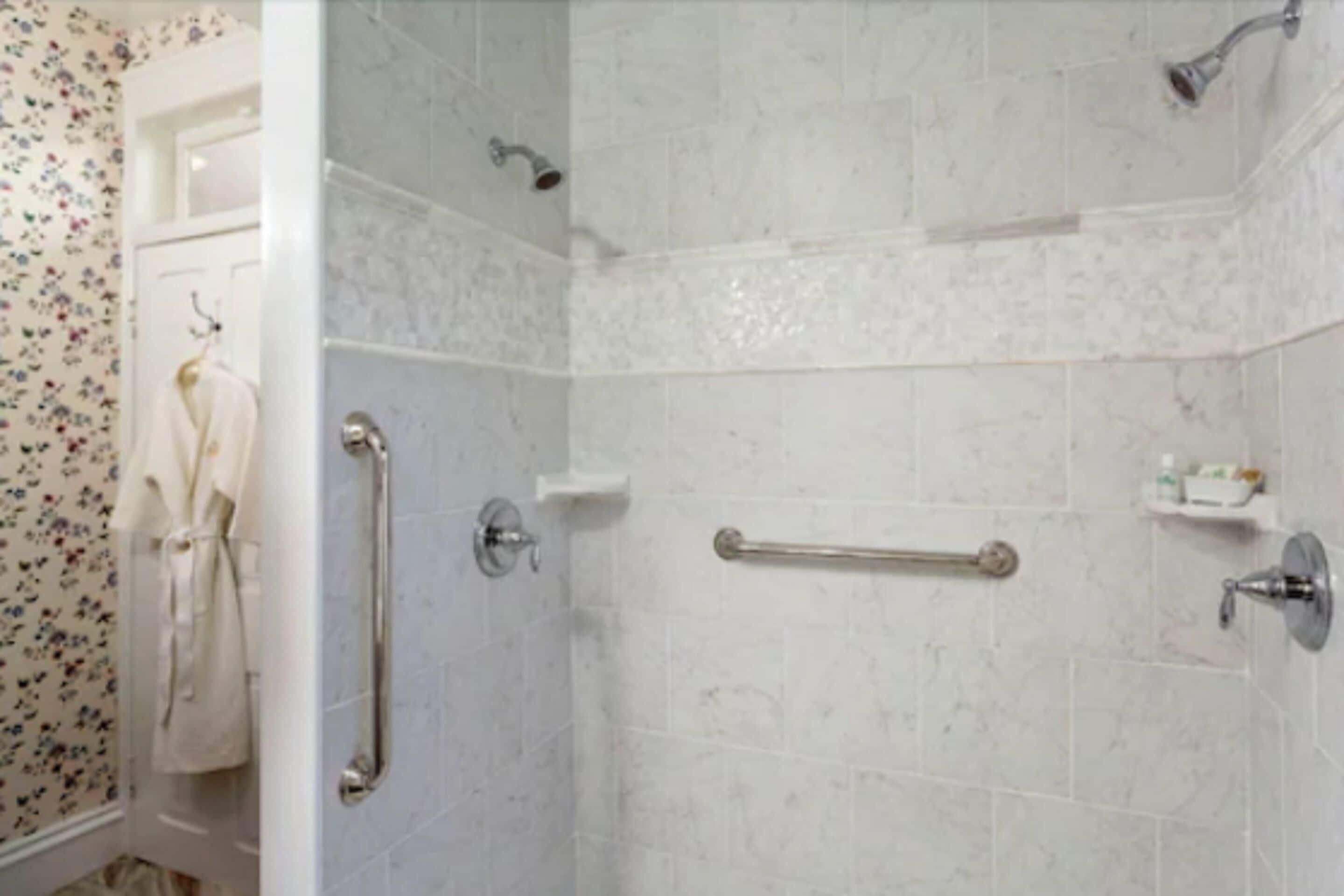 A bathroom with white tiled walls, a shower with a handheld showerhead, a grab bar, and a towel rack with a white towel.