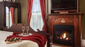 A room with a bed, a fireplace, a television, and a window with red curtains. - The Queen Victoria B&B (Cape May)