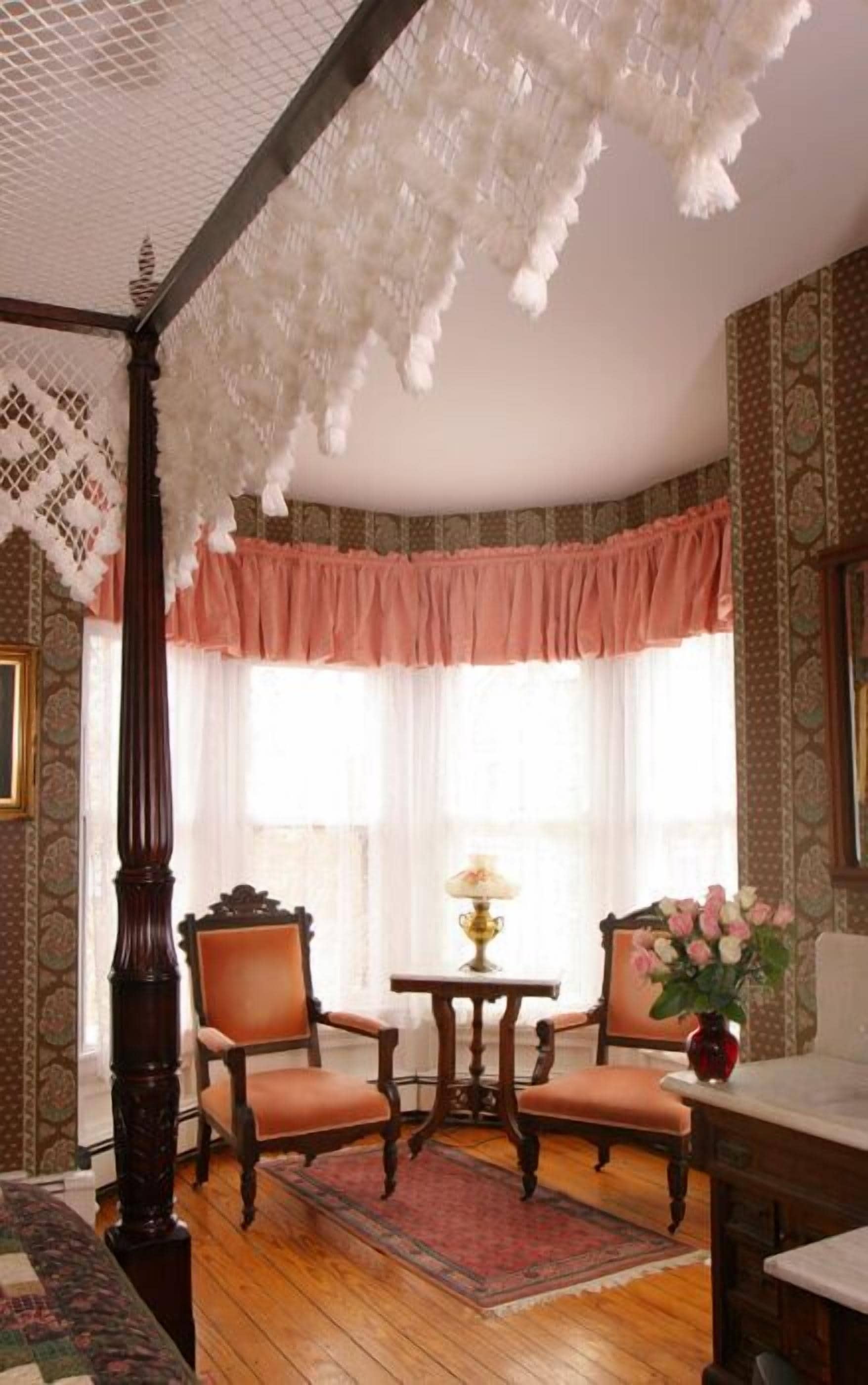 A traditional room with a four-poster bed, two chairs, a table with a lamp, and a vase of flowers.