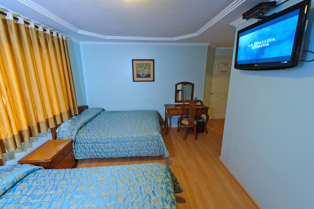 Habitació d'hotel amb dos llits, una taula de nit, una cadira, un televisor i una finestra amb cortines.