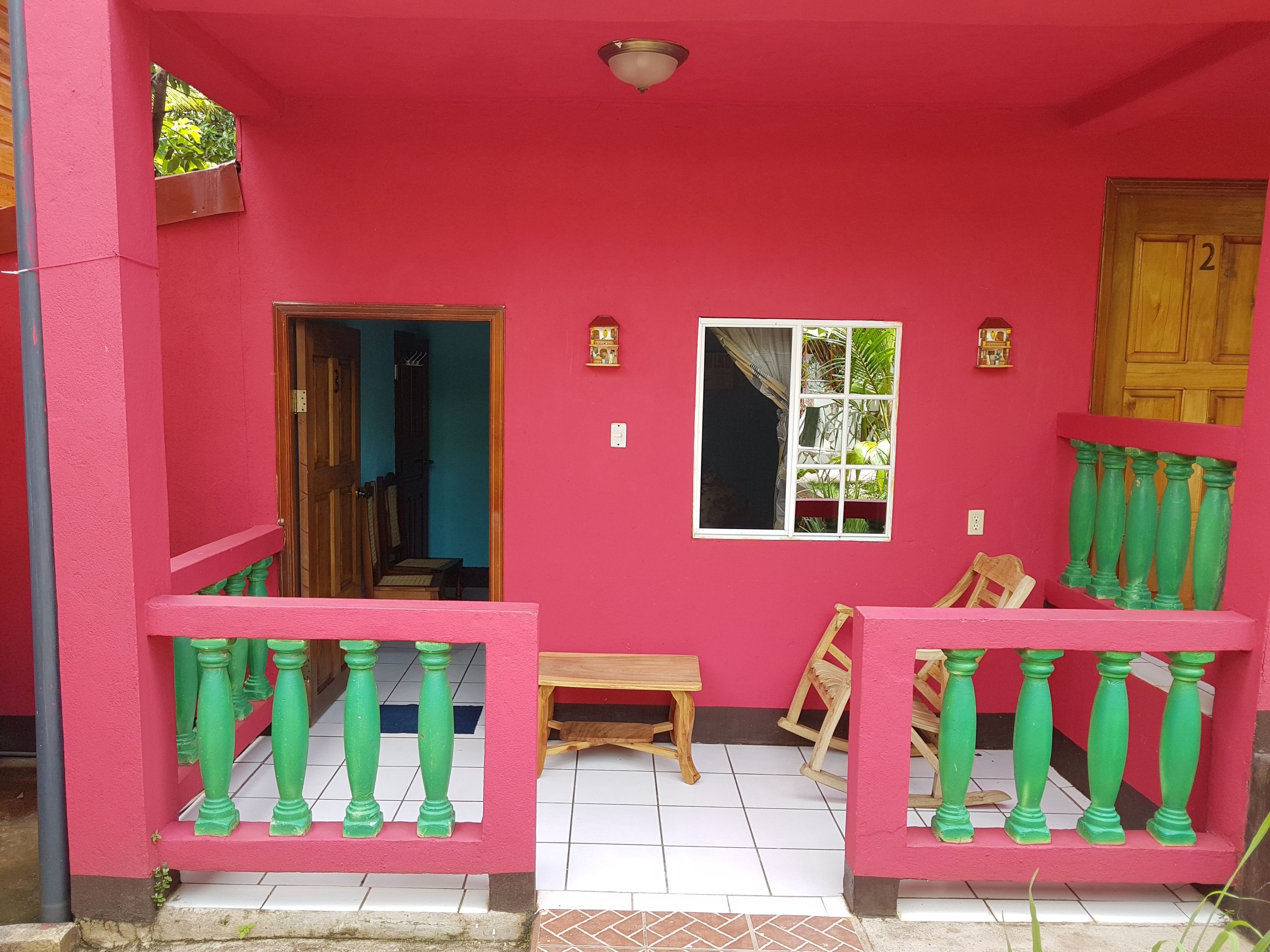 Fachada rosa con puerta de madera marcada con el número '2', ventana con vista a la vegetación y un pequeño banco de madera.