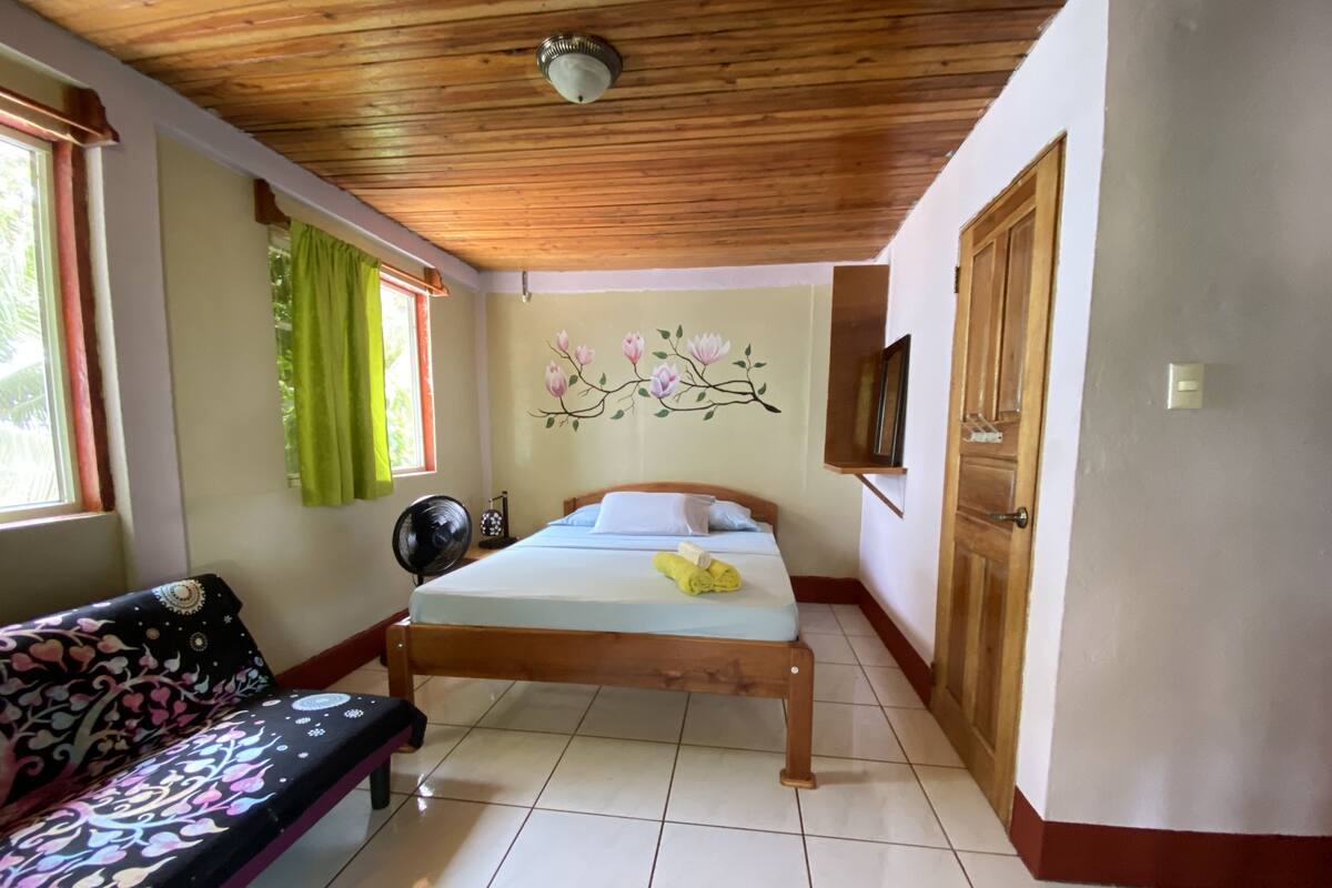 Une chambre à coucher avec un lit en bois, un canapé, un ventilateur, un téléviseur fixé au mur et une porte.