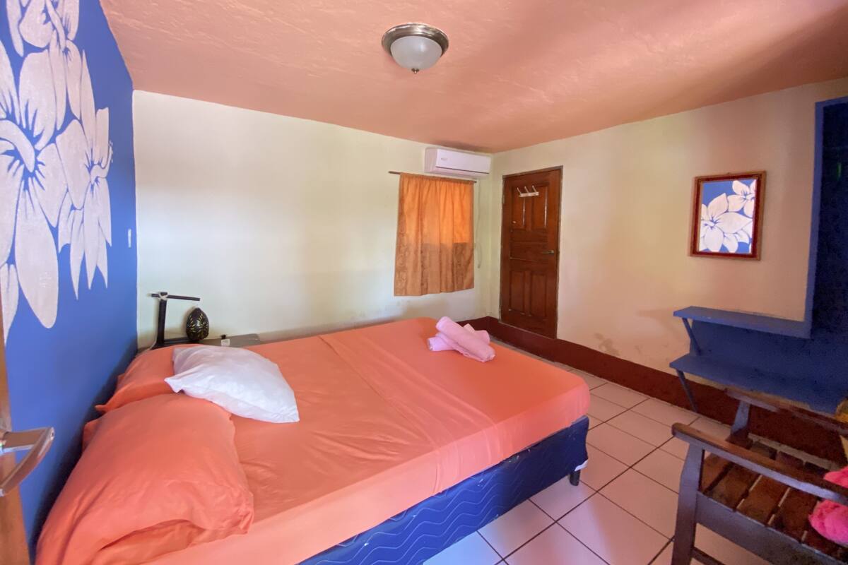 Une chambre à coucher comprenant un lit, un ventilateur de plafond, un appareil de climatisation, une porte en bois et une petite table avec une chaise.