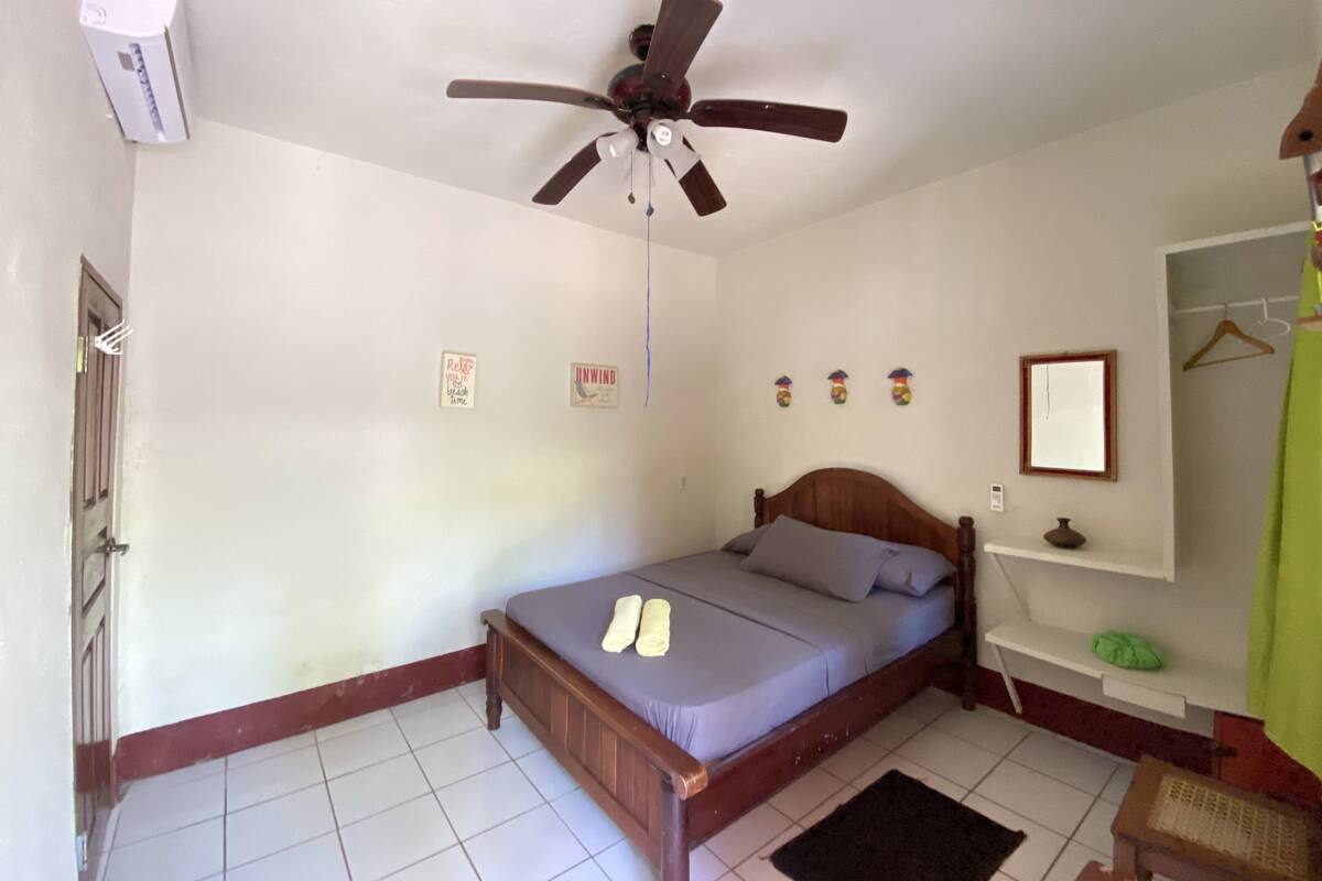 Une chambre à coucher avec un lit en bois, un ventilateur de plafond, un climatiseur blanc, une petite étagère avec un vase, et une porte sur laquelle est accrochée une serviette.