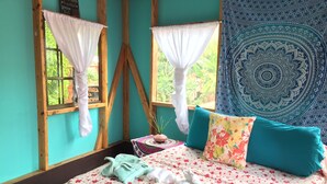 Una habitación con cama, ventana con cortinas, un tapiz y una pequeña mesa con una planta.