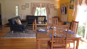 A living room with a sofa, a dining table set with plates and glasses, wooden chairs, a coffee table, and a TV on the wall. - Marzden Cottage (Daylesford)