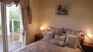 A bedroom with a large bed, floral bedding, two bedside tables with lamps, a painting on the wall, and a view of a garden through a window. - Marzden Cottage (Daylesford)