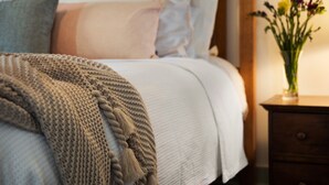 A neatly made bed with a knitted blanket and pillows, a bedside table with a vase of flowers, and a wooden nightstand. - Monigatti (Hepburn Springs)