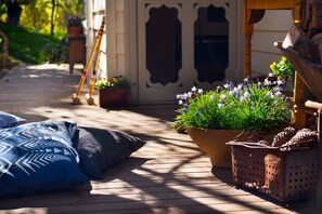 Um deck de madeira com vasos de plantas, uma vassoura e uma almofada azul estampada.
