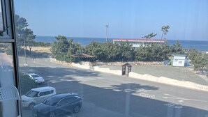 A balcony with white chairs and a small table, overlooking a parking area with cars and a view of the sea and buildings. - Pinebeach Hotel Pohang (Pohang)