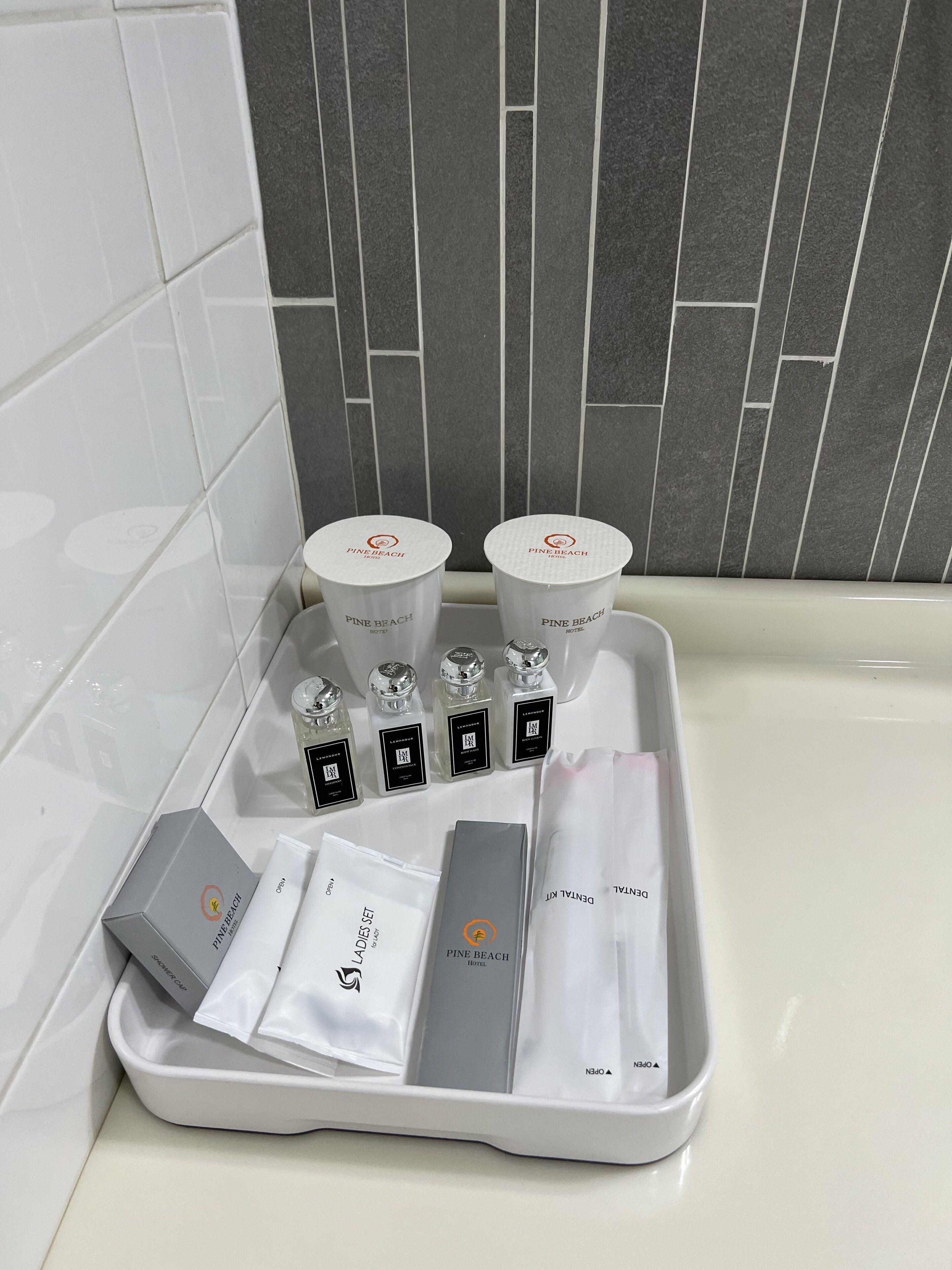 A bathroom vanity with a white sink, a tray holding various toiletries including Pine Beach Hotel branded items, and a grey tiled wall in the background.