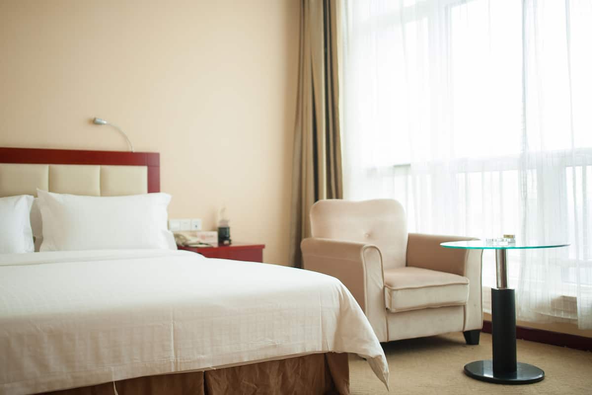 Quarto de hotel com uma cama grande, uma cadeira, uma mesinha e uma janela com cortinas.