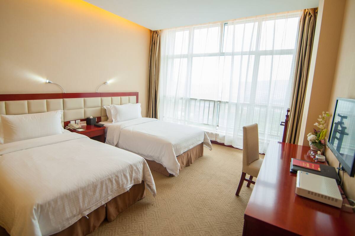 Quarto de hotel com duas camas, uma escrivaninha, uma cadeira e uma janela grande com cortinas.