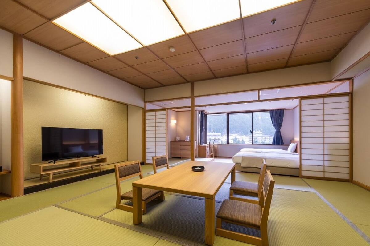 A traditional Japanese-style room with tatami flooring, a television, a dining table with chairs, and a bed with a view of the city.