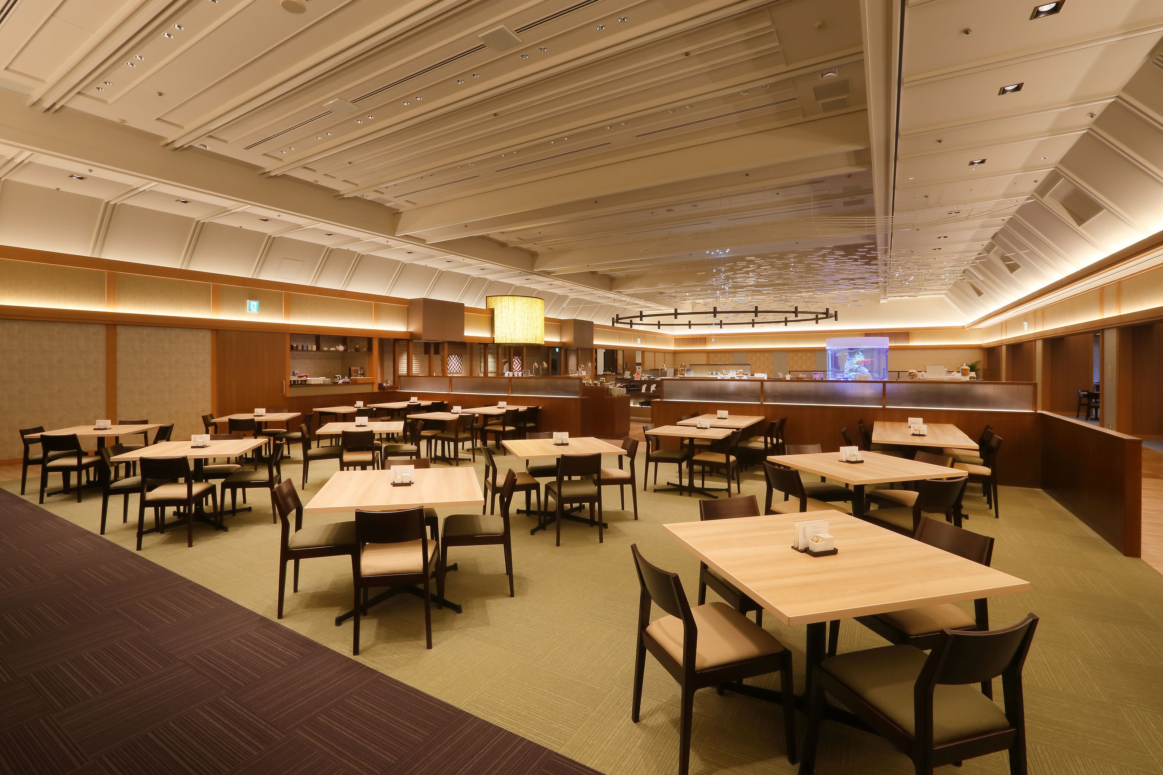 Un amplio salón comedor de hotel con varias mesas y sillas, una zona de bar central y una pantalla grande mostrando contenido.