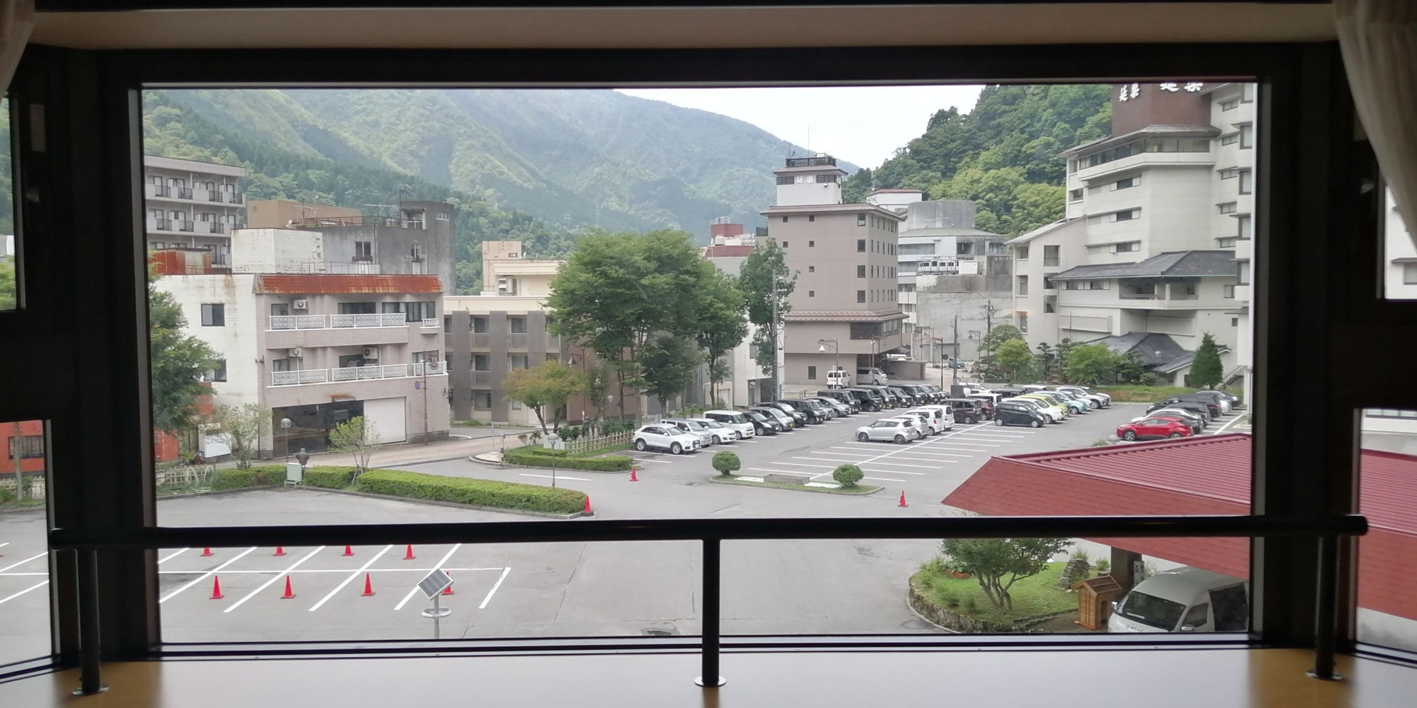 A view from a window overlooking a parking lot with multiple cars, a red-roofed building, and surrounding residential buildings with a mountainous backdrop.