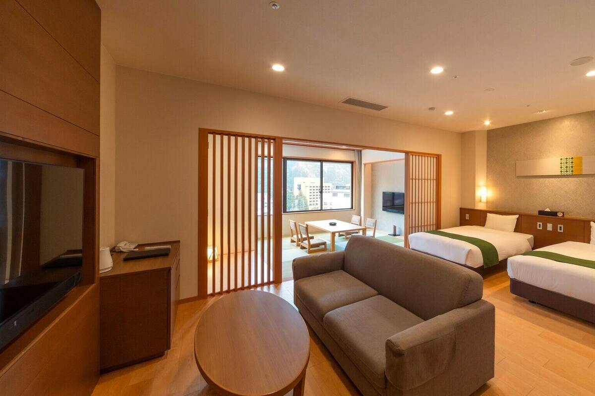 A hotel room with two beds, a sofa, a TV, a desk, and a sliding door leading to a balcony.