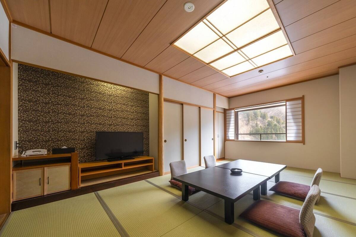 A traditional Japanese-style room with tatami flooring, a low table with cushions, a television on a wooden stand, and a window with a view of trees.