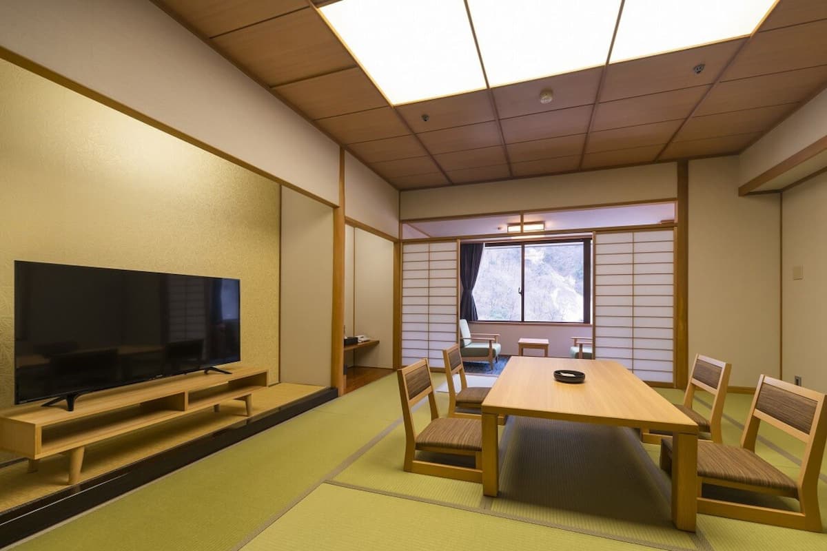 A traditional Japanese-style room with tatami flooring, a television on a low stand, a wooden table with chairs, and a window with a view of trees.