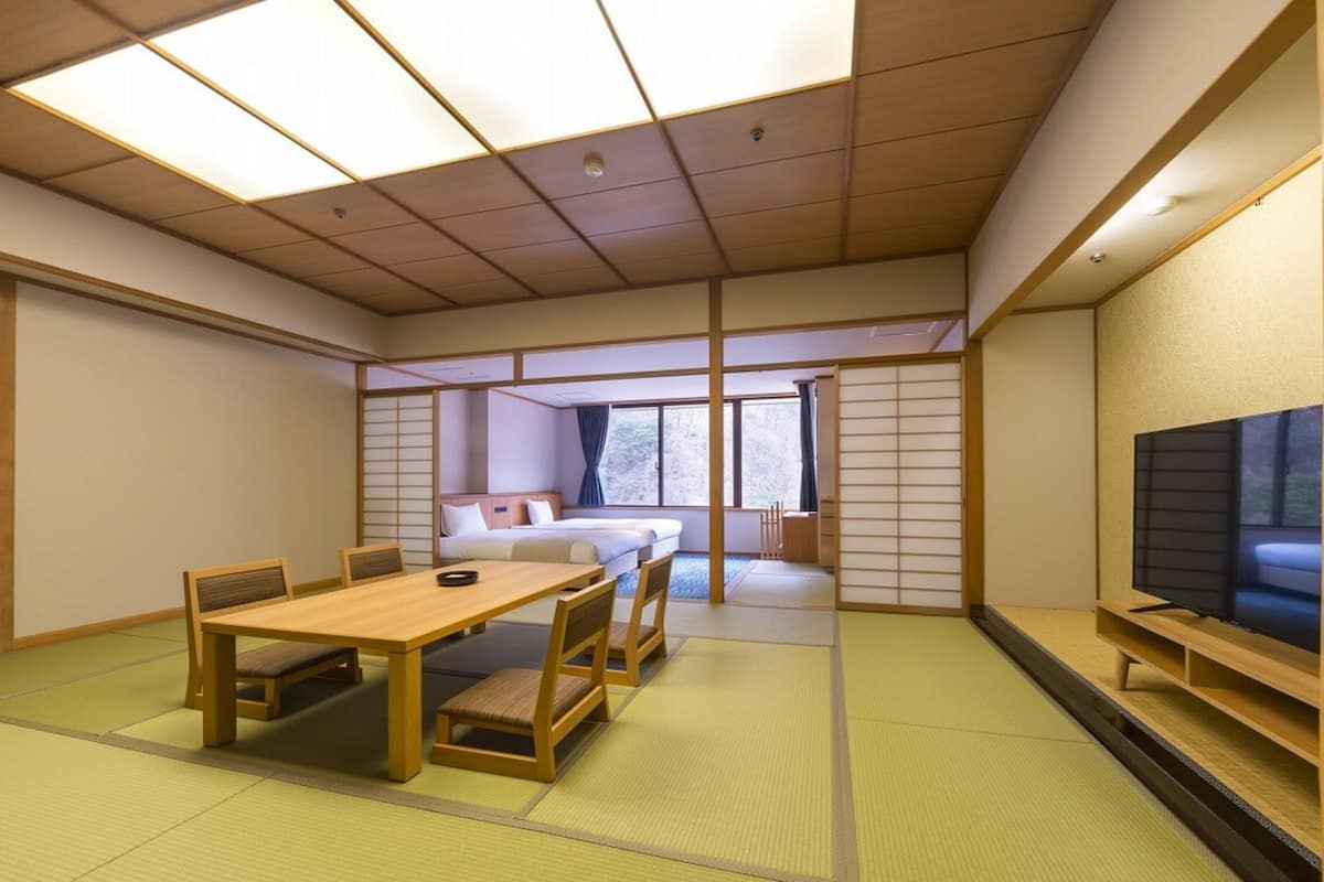 A traditional Japanese-style room with tatami flooring, a low table with chairs, a bed with a view to the outside, and a television on a shelf.