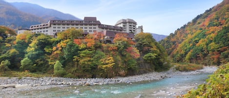 A hotel with multiple floors, a river in the foreground, and a mountainous backdrop.
